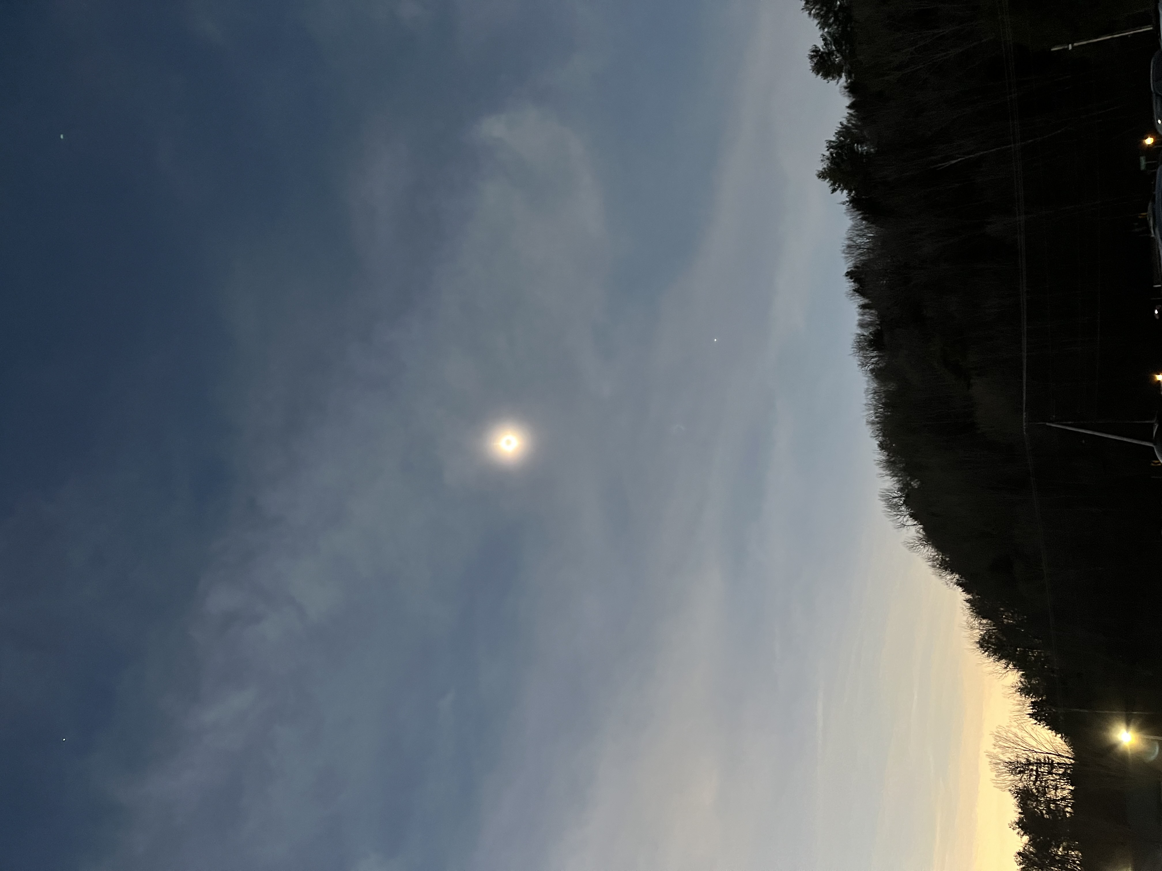 Sun during totality above the silhouette of a forested hill. Several other celestial bodies are visible in the sky around it
